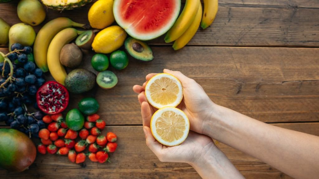 Hands holding sliced lemon surrounded by an assortment of fresh fruits on a wooden table.